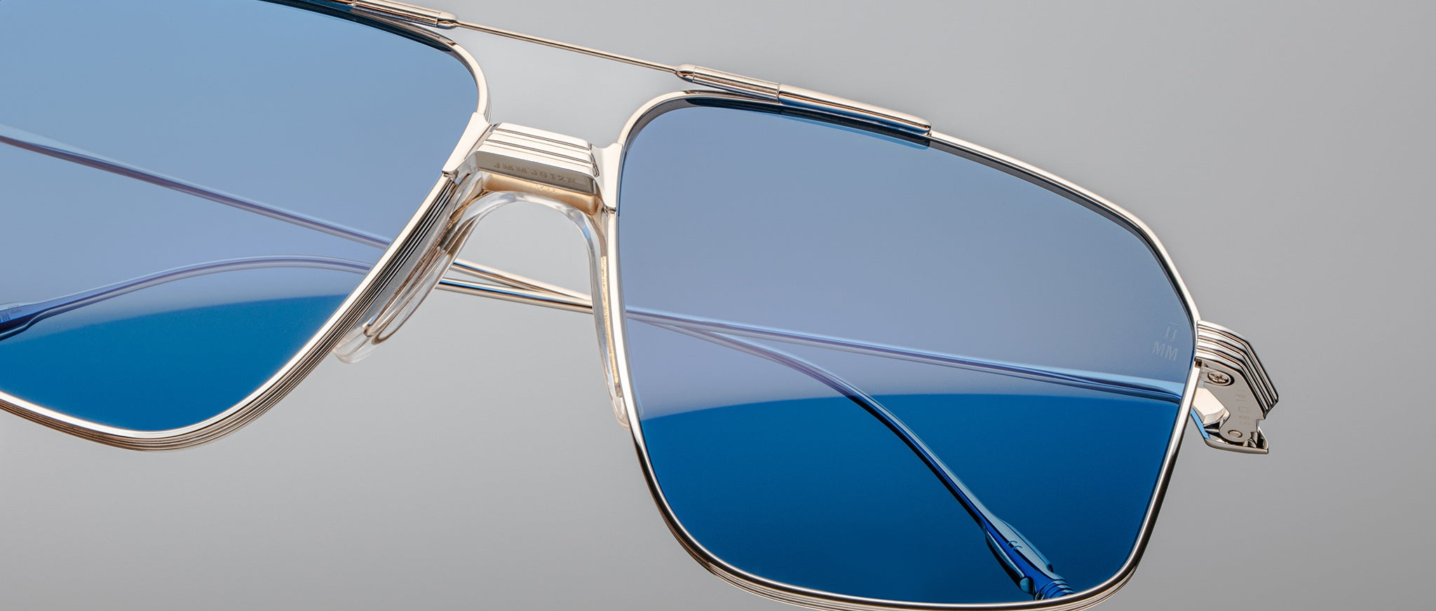 Close-up of Jacques Marie Mage Jagger oversized aviator sunglasses by Jacques Marie Mage, featuring gold metal frames, blue-tinted lenses, a double bridge, and lightweight beta titanium construction, shown against a plain grey background.