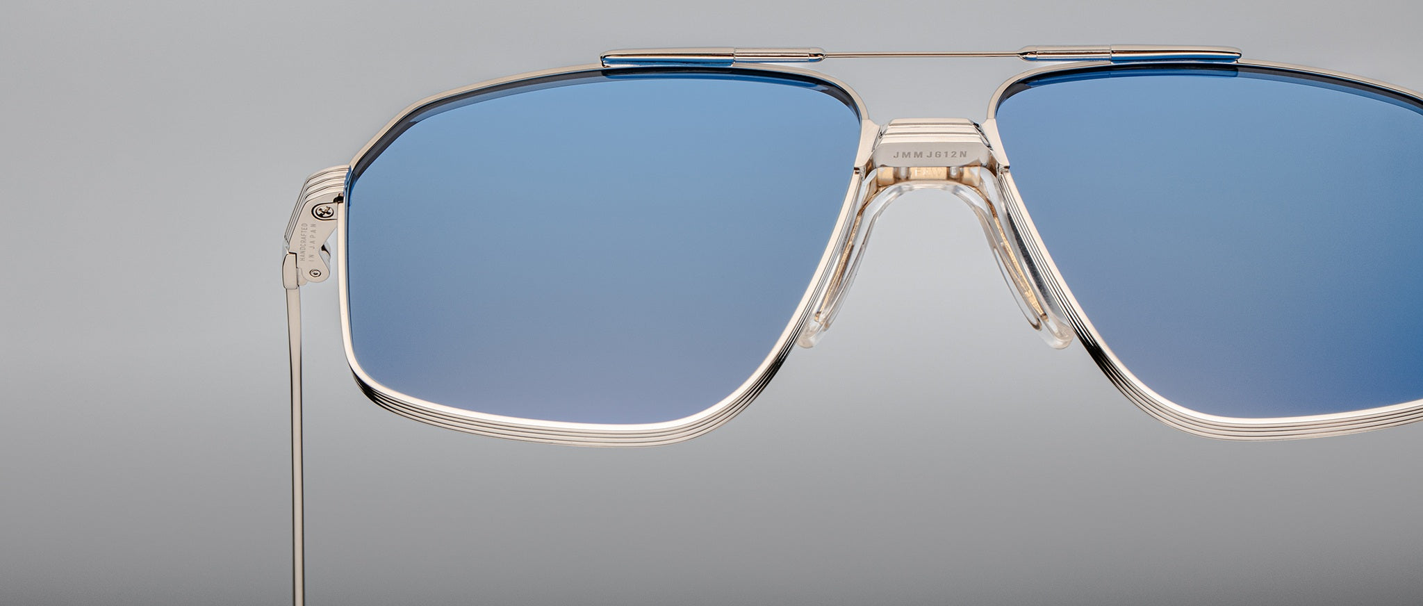 Close-up of Jacques Marie Mage Jagger sunglasses by Jacques Marie Mage, featuring oversized aviator design, blue tinted lenses, and thin beta titanium frames set against a plain grey background.