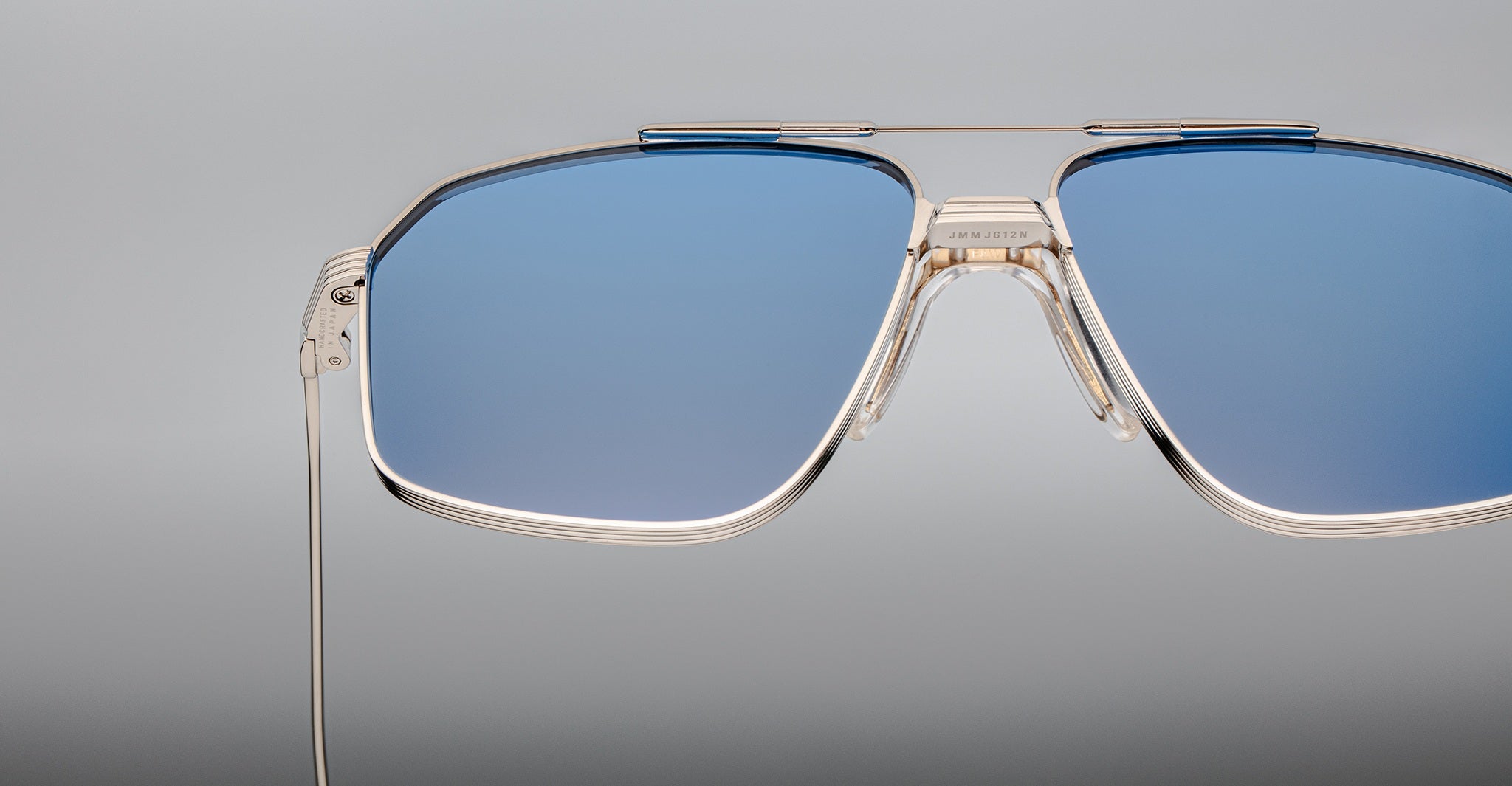 Close-up of Jacques Marie Mage Jagger sunglasses by Jacques Marie Mage, featuring oversized aviator design, blue tinted lenses, and thin beta titanium frames set against a plain grey background.