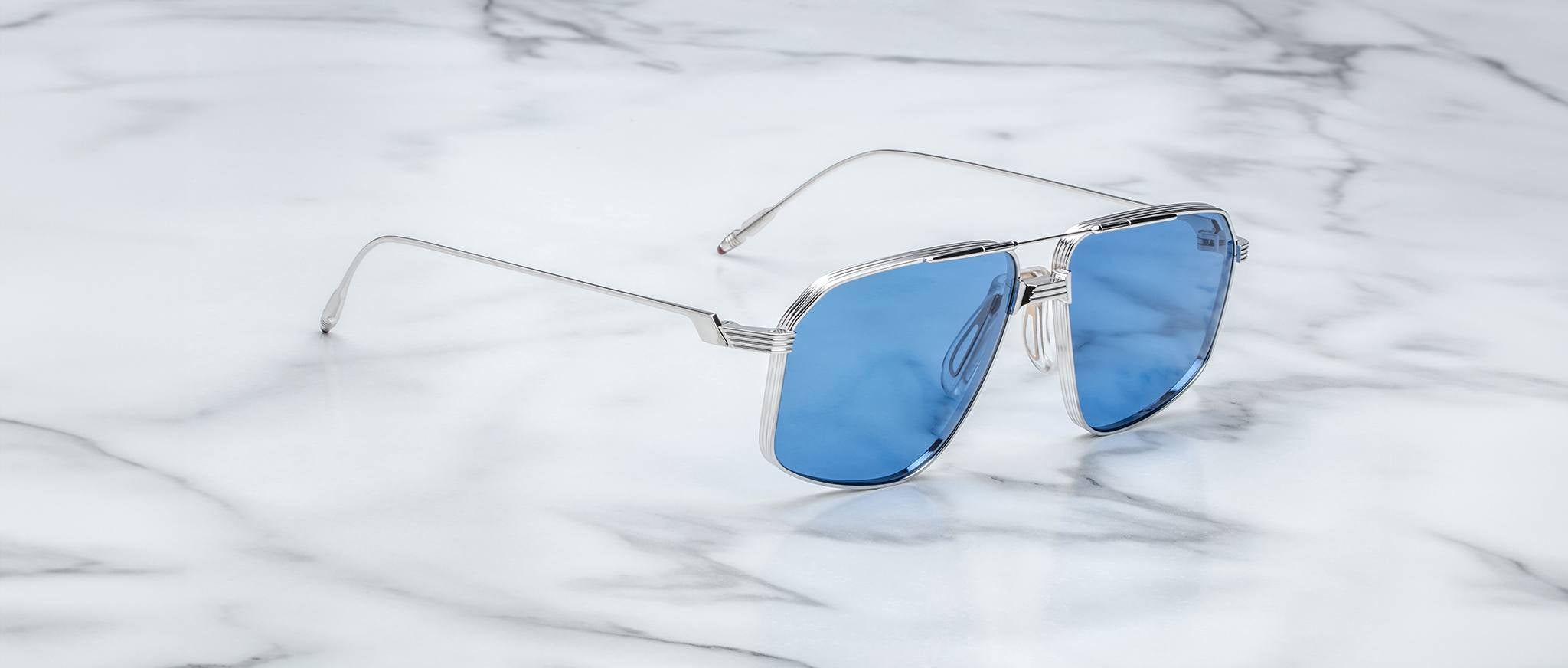 A pair of Jacques Marie Mage Jagger aviator sunglasses with silver frames and blue lenses rests on white marble, highlighting their sleek beta titanium design.