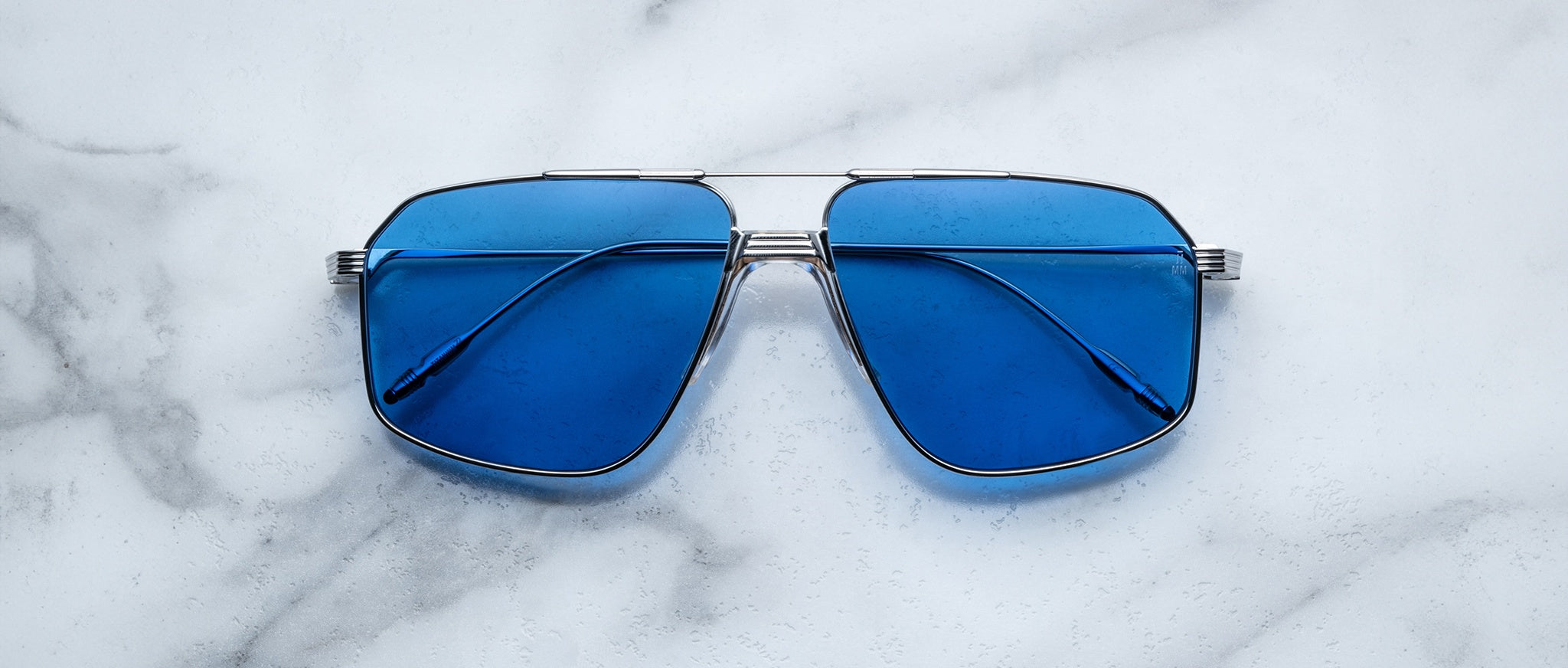 The Jacques Marie Mage Jagger sunglasses by Jacques Marie Mage feature oversized aviator frames in silver beta titanium with blue-tinted lenses, displayed on a white marble surface.
