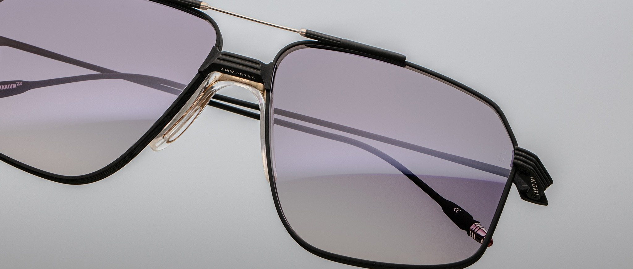 Close-up of Jacques Marie Mage Jagger oversized aviator sunglasses by Jacques Marie Mage, with a thin black and gold beta titanium frame and light purple tinted lenses, set against a plain light background.
