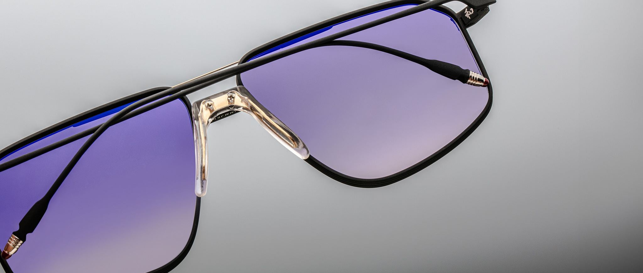 Close-up of Jacques Marie Mage Jagger sunglasses by Jacques Marie Mage, with oversized black frames, beta titanium accents, and gradient purple lenses on a light grey background.