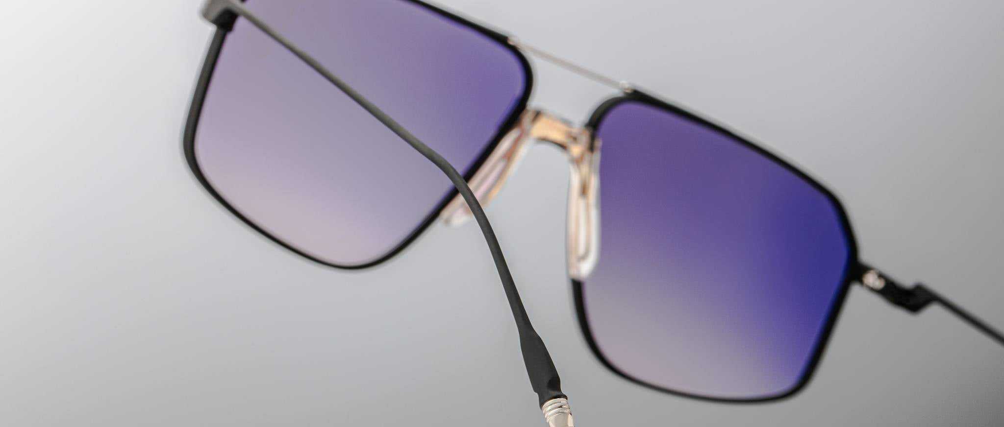 Close-up of Jacques Marie Mage Jagger sunglasses featuring oversized square purple-tinted lenses, black frames, and gold accents in premium beta titanium by Jacques Marie Mage, displayed against a neutral background.