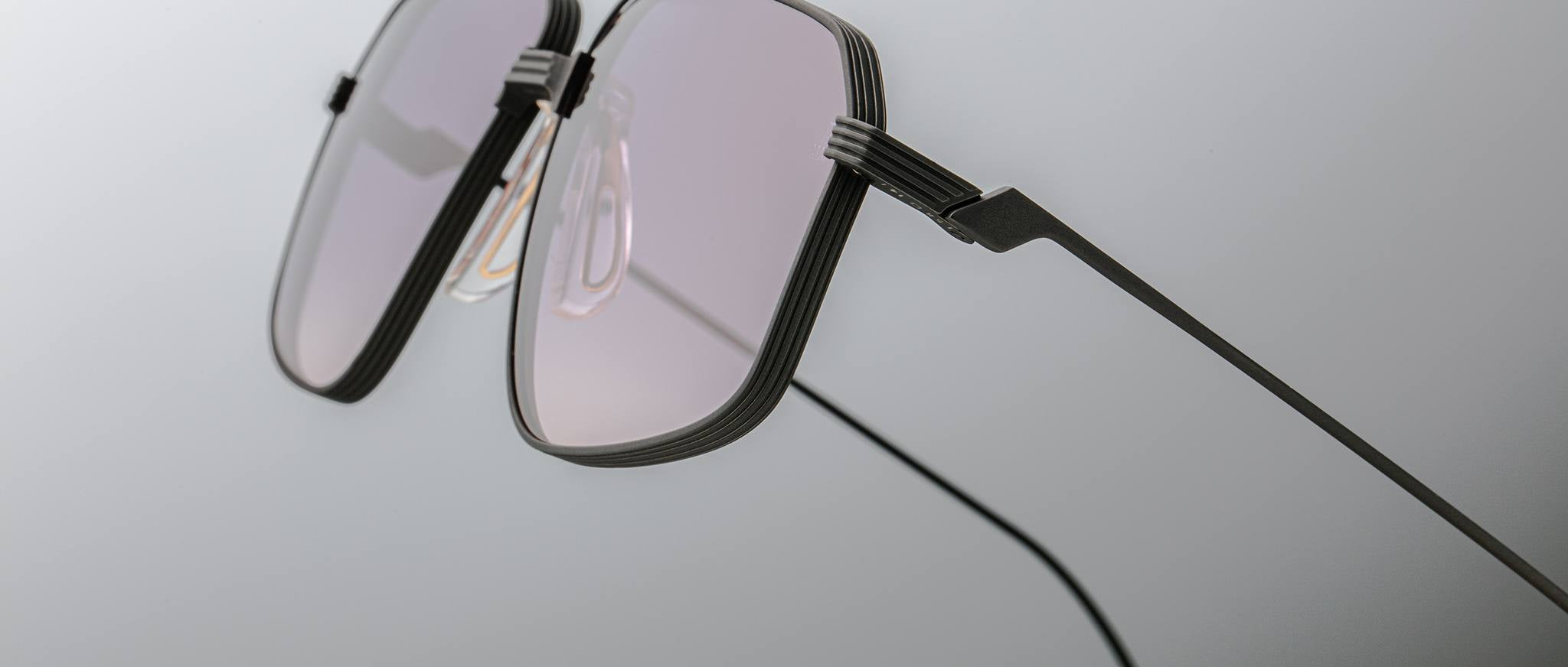 Close-up of Jacques Marie Mage Jagger eyeglasses by Jacques Marie Mage, featuring thin black beta titanium frames and lightly tinted lenses set against a plain, light grey background.