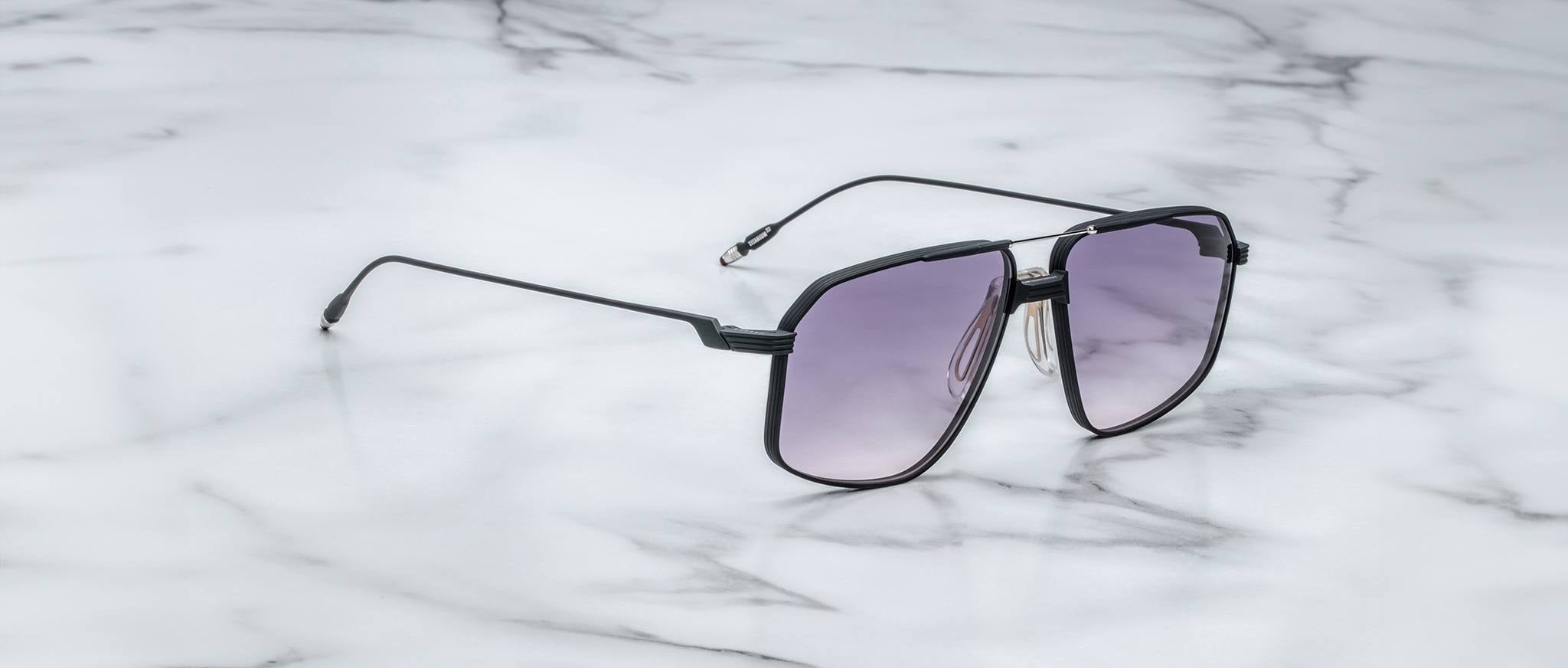 The Jacques Marie Mage Jagger, a pair of oversized aviator sunglasses by Jacques Marie Mage with black frames and gradient lenses, rests on a white marble surface.