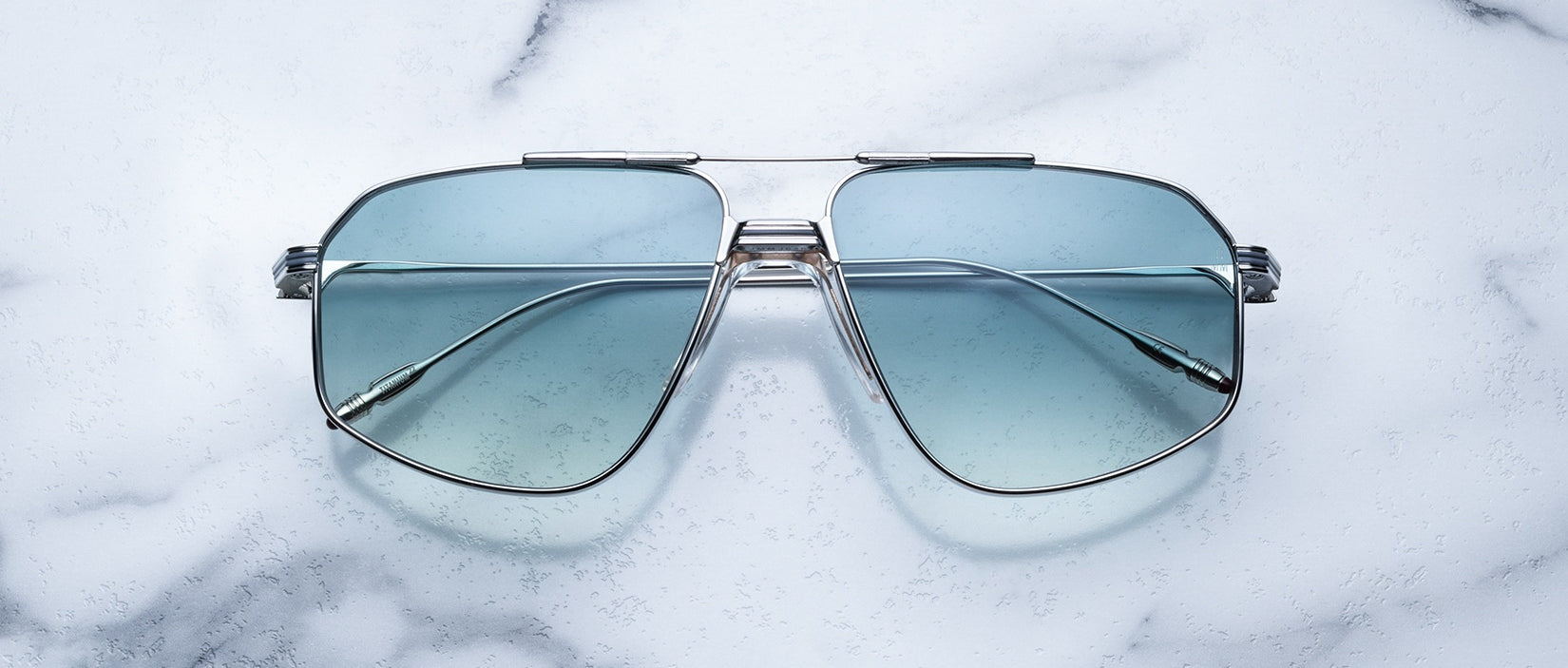 Jacques Marie Mage Jagger sunglasses by Jacques Marie Mage, featuring blue-tinted oversized aviator lenses, are displayed elegantly on a white marble surface.