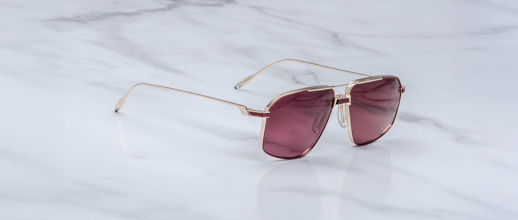 A pair of Jacques Marie Mage Jagger sunglasses, featuring oversized gold frames and large square maroon-tinted lenses, rests on a white marble surface.