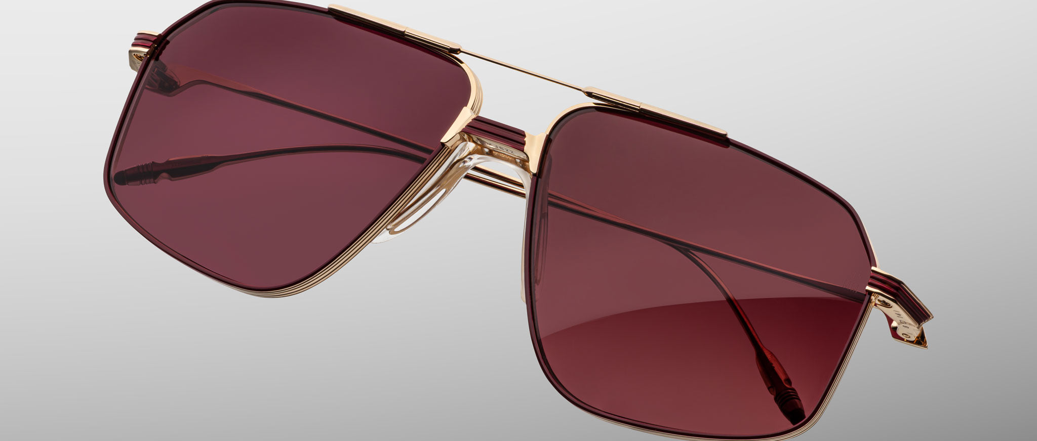 The Jacques Marie Mage Jagger sunglasses by Jacques Marie Mage feature a gold frame and large square burgundy-tinted lenses, showcased against a plain gray background.