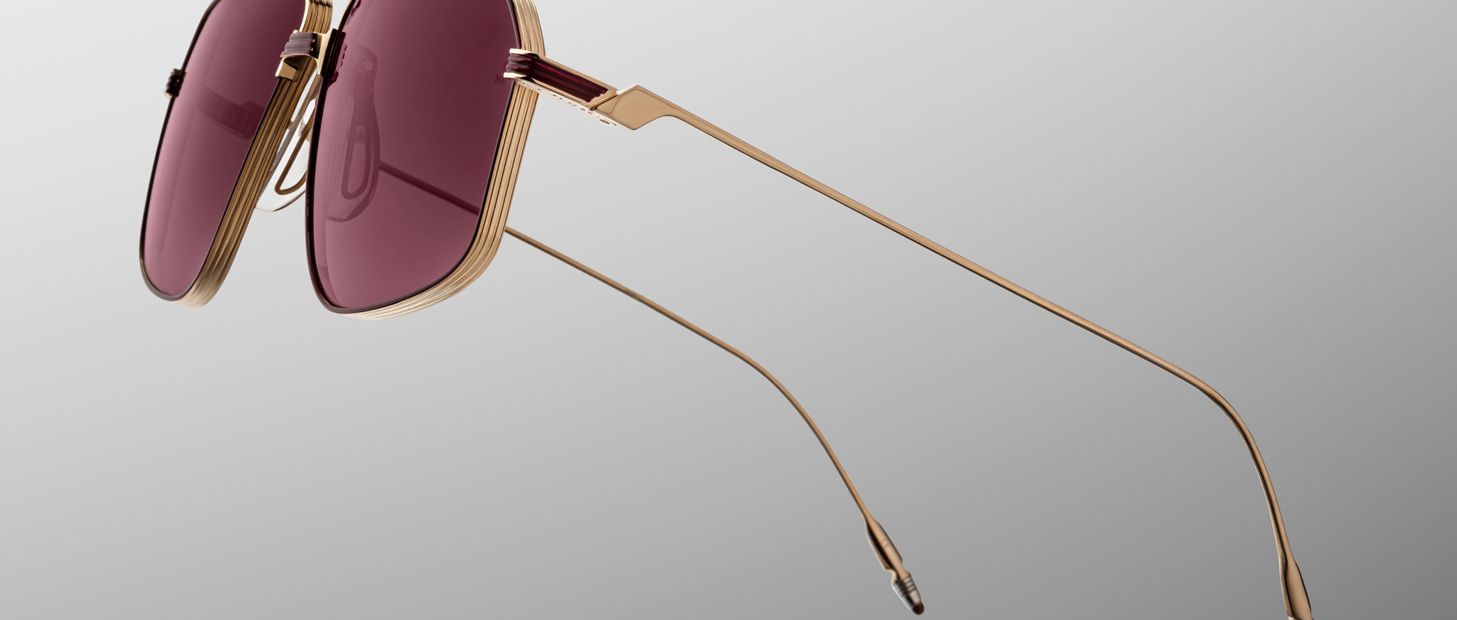 The Jacques Marie Mage Jagger by Jacques Marie Mage features gold-framed oversized aviators with large, rectangular burgundy-tinted beta titanium lenses, inspired by iconic designs and displayed on a plain gradient background.
