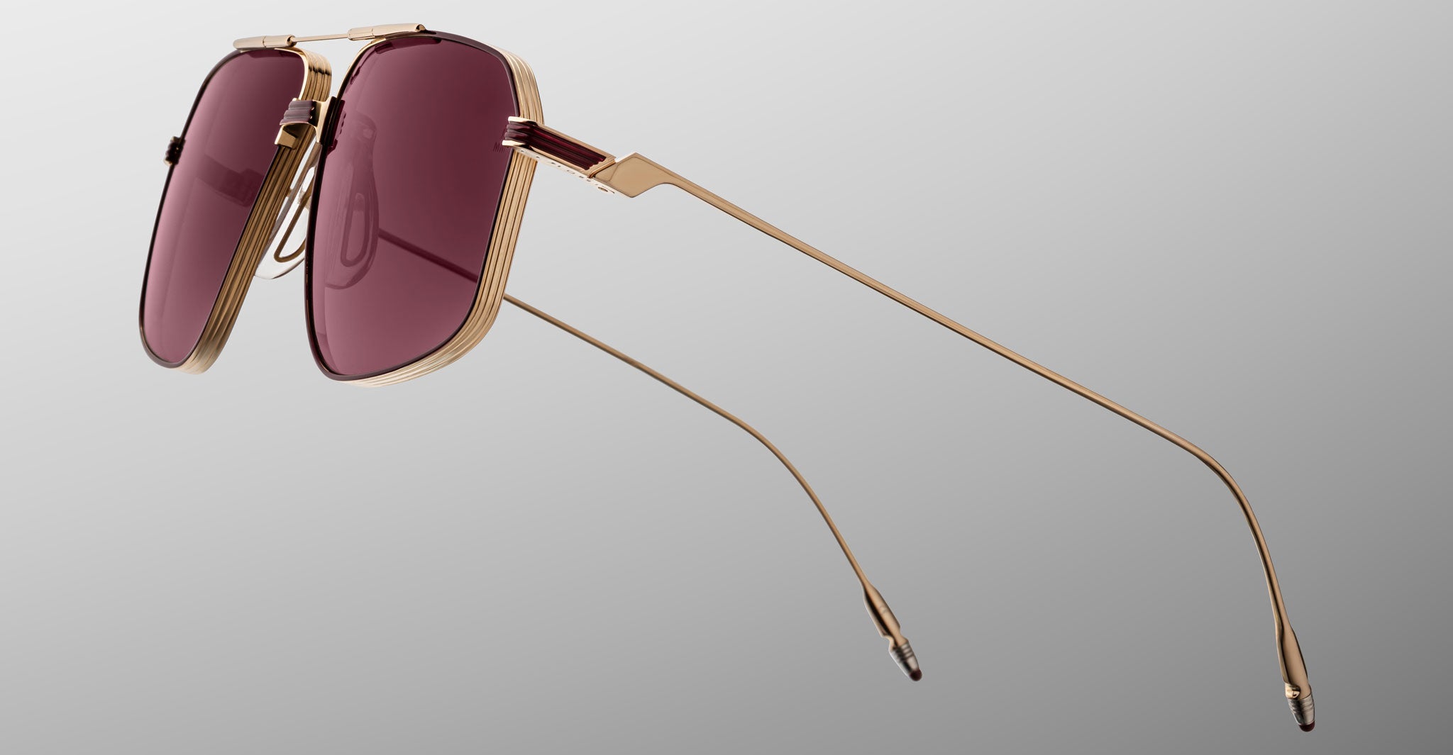The Jacques Marie Mage Jagger by Jacques Marie Mage features gold-framed oversized aviators with large, rectangular burgundy-tinted beta titanium lenses, inspired by iconic designs and displayed on a plain gradient background.