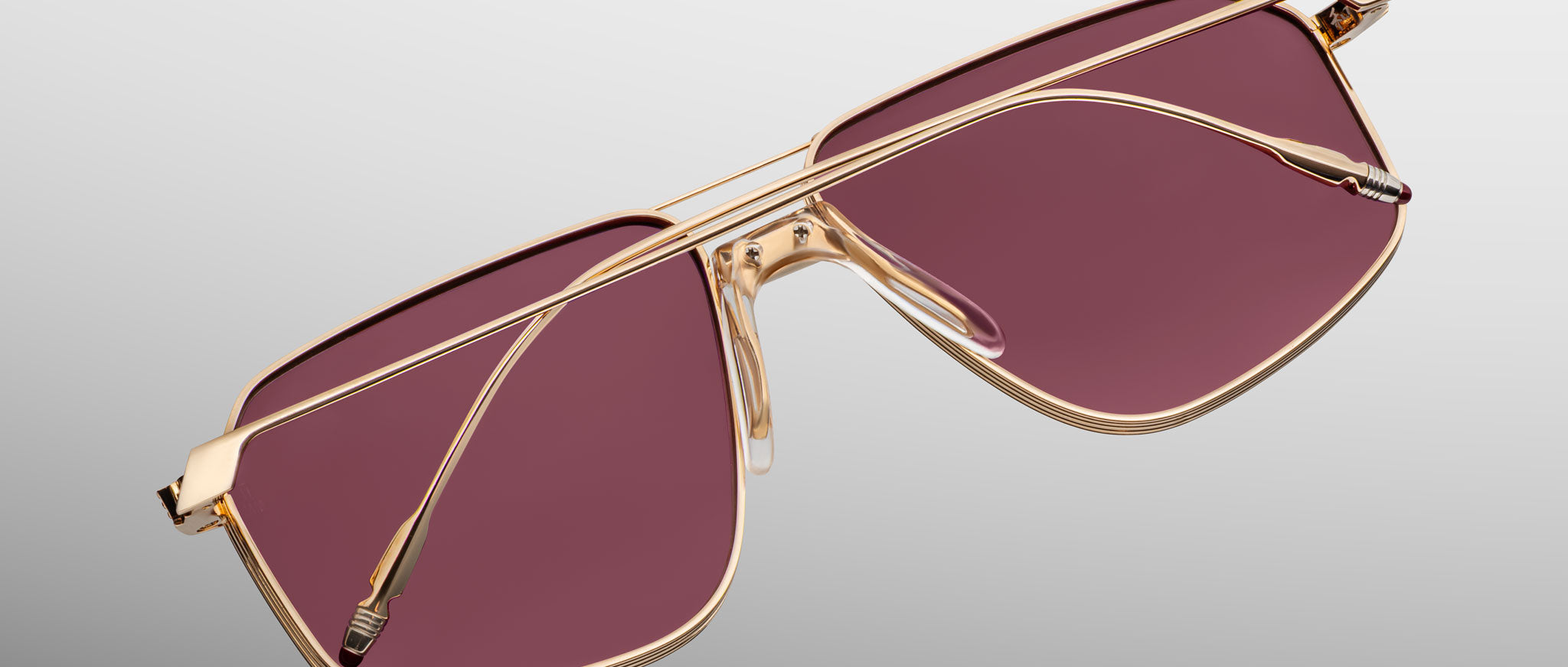 Jacques Marie Mage Jagger sunglasses by Jacques Marie Mage feature a beta titanium frame, gold rims, oversized rectangular rose-tinted lenses, and are shown on a light gray background.