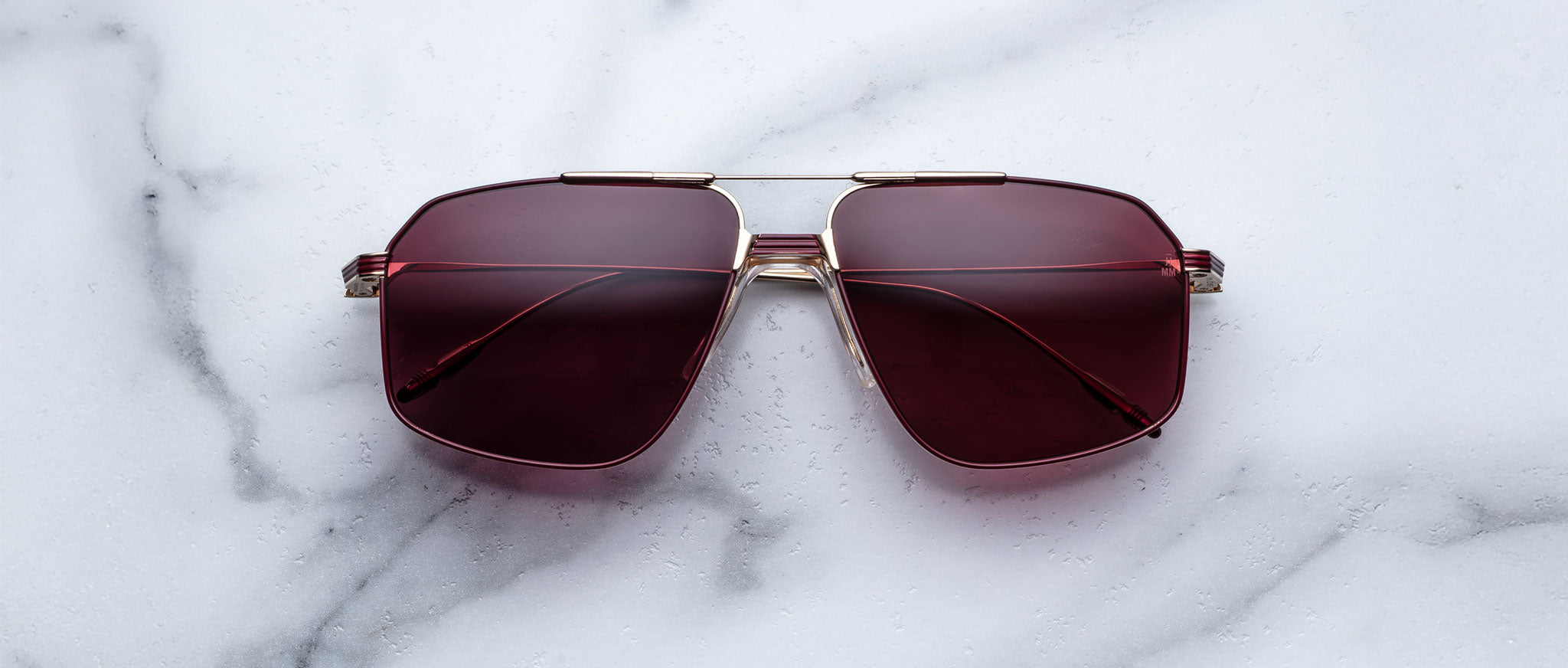 The Jacques Marie Mage Jagger sunglasses by Jacques Marie Mage feature oversized aviator frames in gold with dark red-tinted lenses, displayed elegantly on a white marble surface.