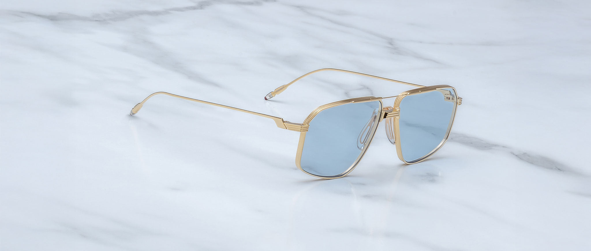 The Jacques Marie Mage Jagger sunglasses by Jacques Marie Mage, featuring gold oversized aviator frames and blue-tinted lenses, are displayed on a white marble surface.
