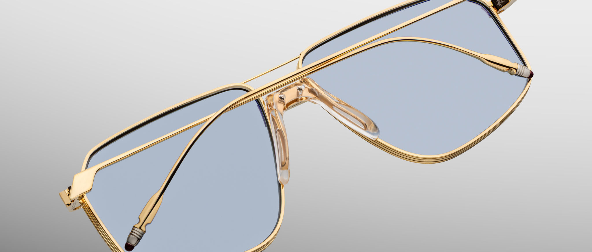The Jacques Marie Mage Jagger sunglasses feature gold beta titanium frames in an oversized aviator shape with clear blue-tinted lenses, showcased against a gradient gray background to capture their bold essence.