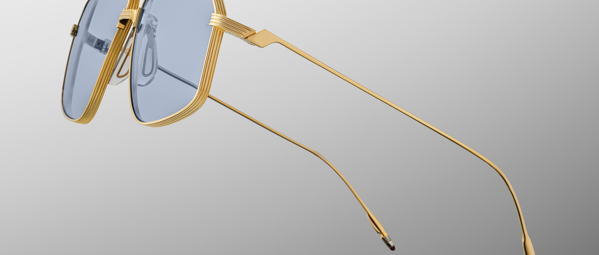 The Jacques Marie Mage Jagger sunglasses by Jacques Marie Mage feature gold beta titanium frames and oversized blue-tinted square lenses, inspired by the Jagger style, displayed on a gray gradient background.