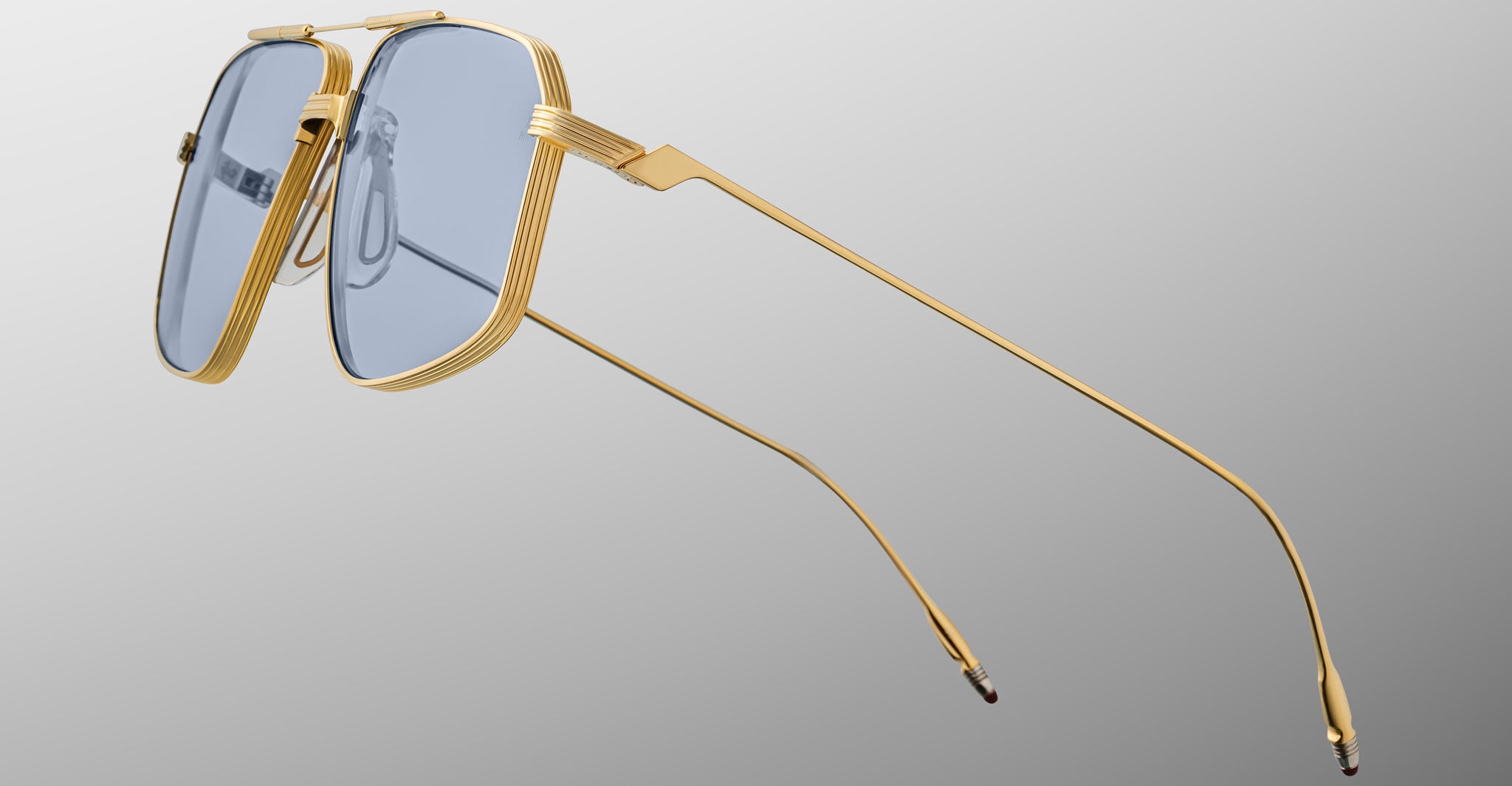 The Jacques Marie Mage Jagger sunglasses by Jacques Marie Mage feature gold beta titanium frames and oversized blue-tinted square lenses, inspired by the Jagger style, displayed on a gray gradient background.