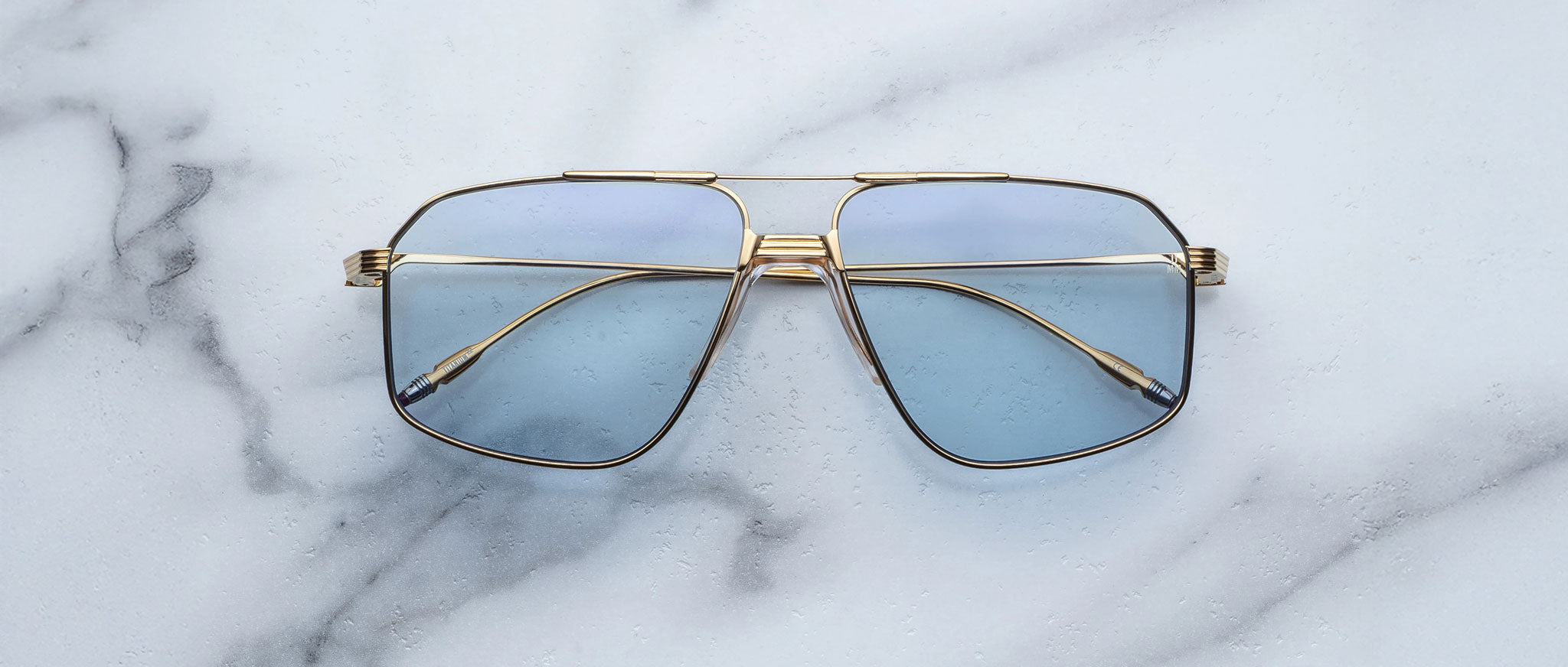 A pair of Jacques Marie Mage Jagger oversized aviator sunglasses with gold frames and blue-tinted lenses by Jacques Marie Mage sits on a white marble surface.