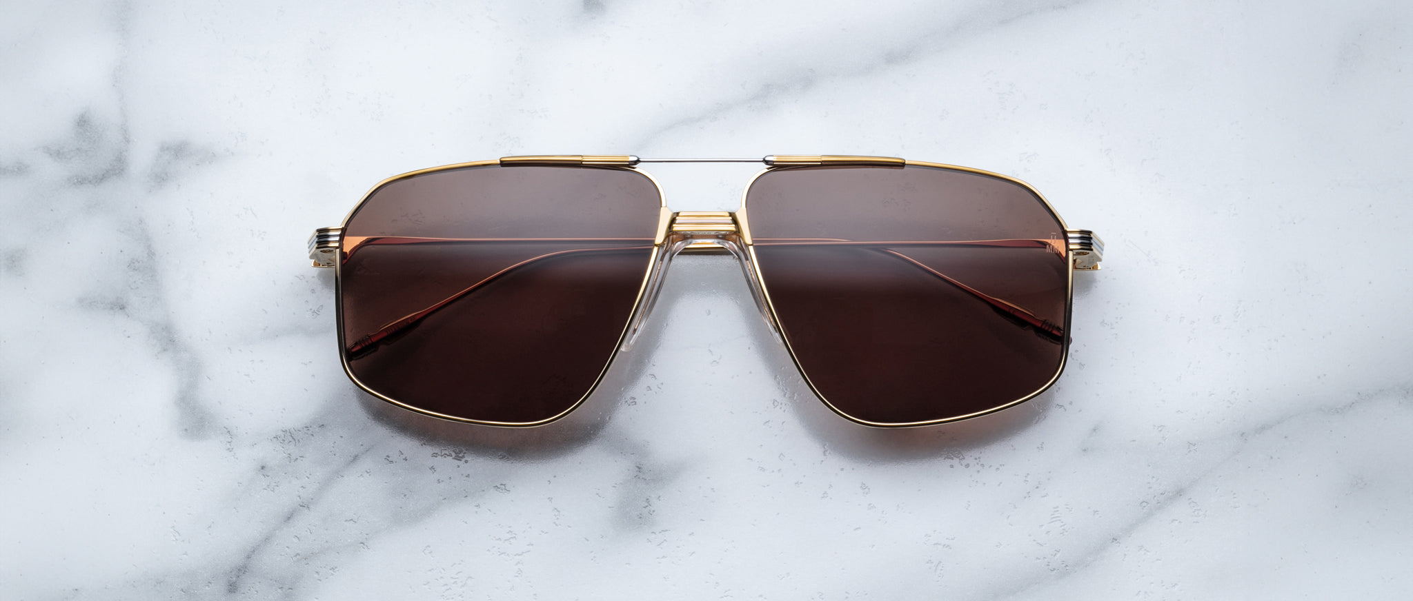 Jacques Marie Mage Jagger sunglasses by Jacques Marie Mage feature gold frames and brown-tinted oversized aviator lenses, displayed on a white marble surface.