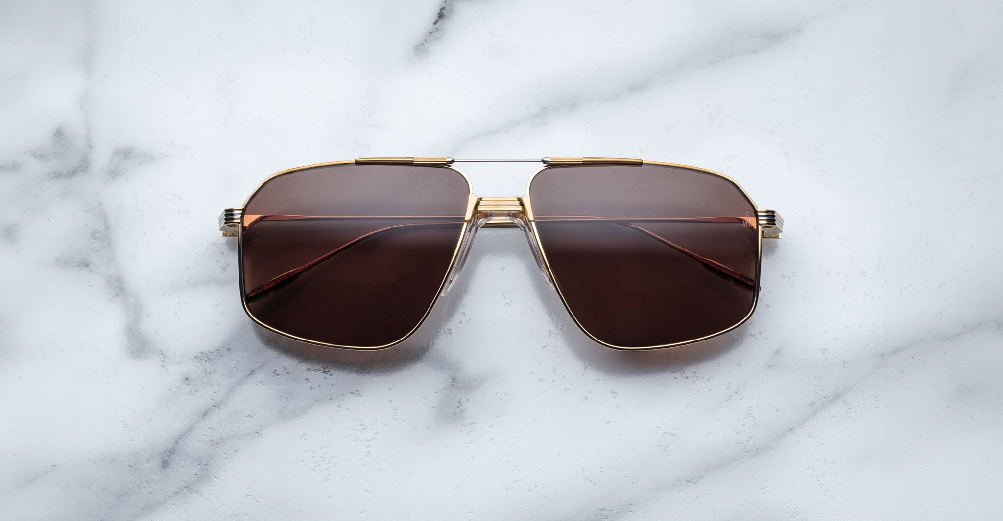 Jacques Marie Mage Jagger sunglasses by Jacques Marie Mage feature gold frames and brown-tinted oversized aviator lenses, displayed on a white marble surface.