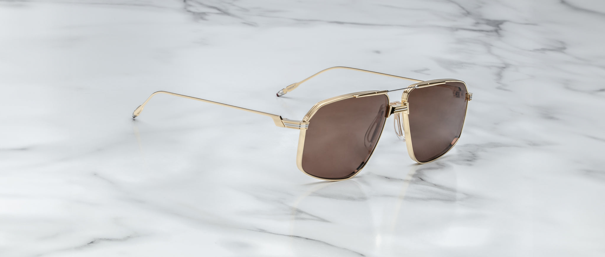 Jacques Marie Mage Jagger gold-framed oversized aviator sunglasses with brown lenses by Jacques Marie Mage rest elegantly on a white marble surface with grey veining.