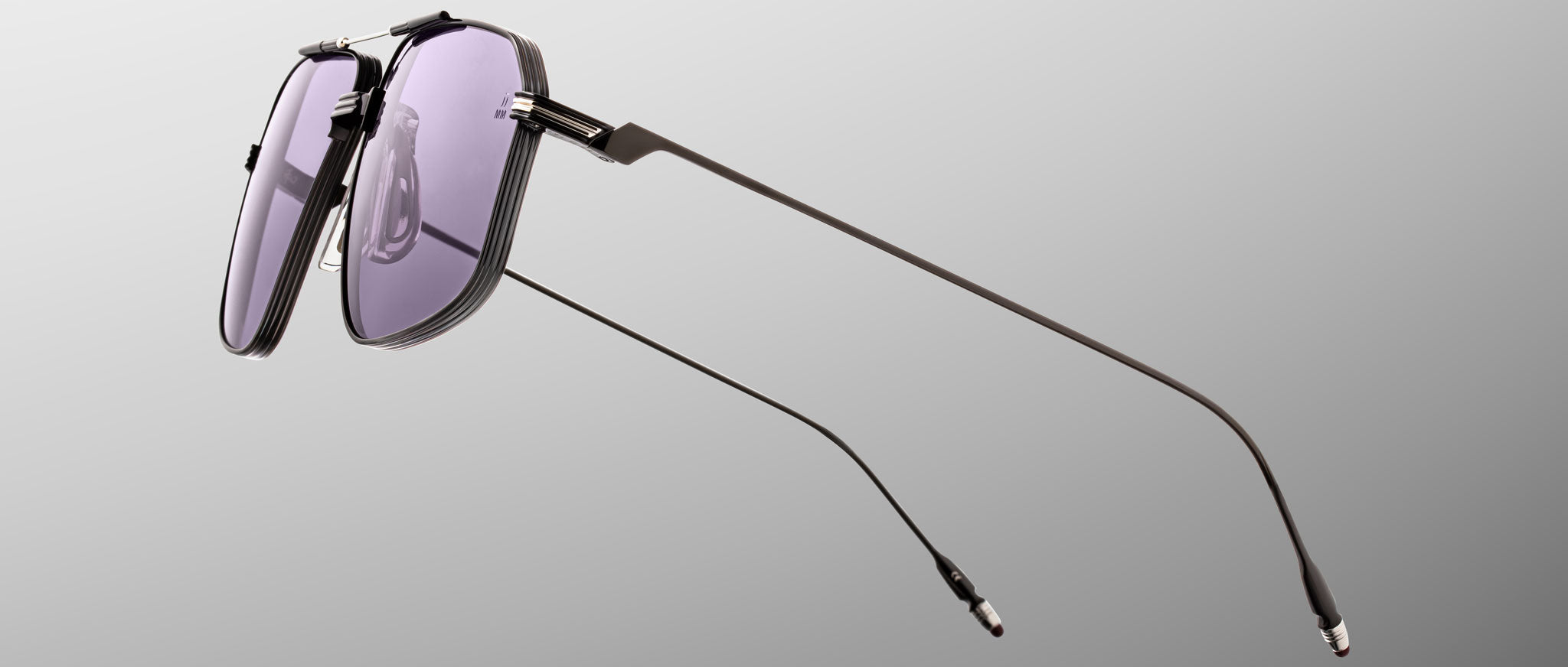 The Jacques Marie Mage Jagger sunglasses by Jacques Marie Mage feature modern oversized aviator frames in thin black beta titanium with purple-tinted lenses, displayed against a plain gray background.