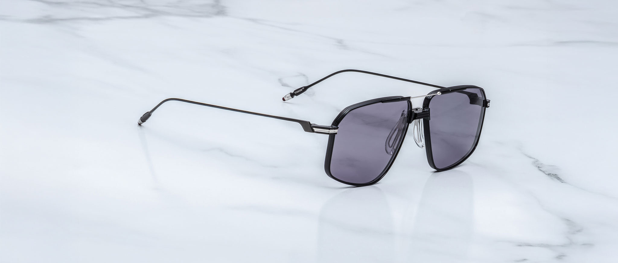 A pair of Jacques Marie Mage Jagger sunglasses by Jacques Marie Mage, featuring an oversized aviator shape with dark lenses and beta titanium accents, rests on a white marble surface.