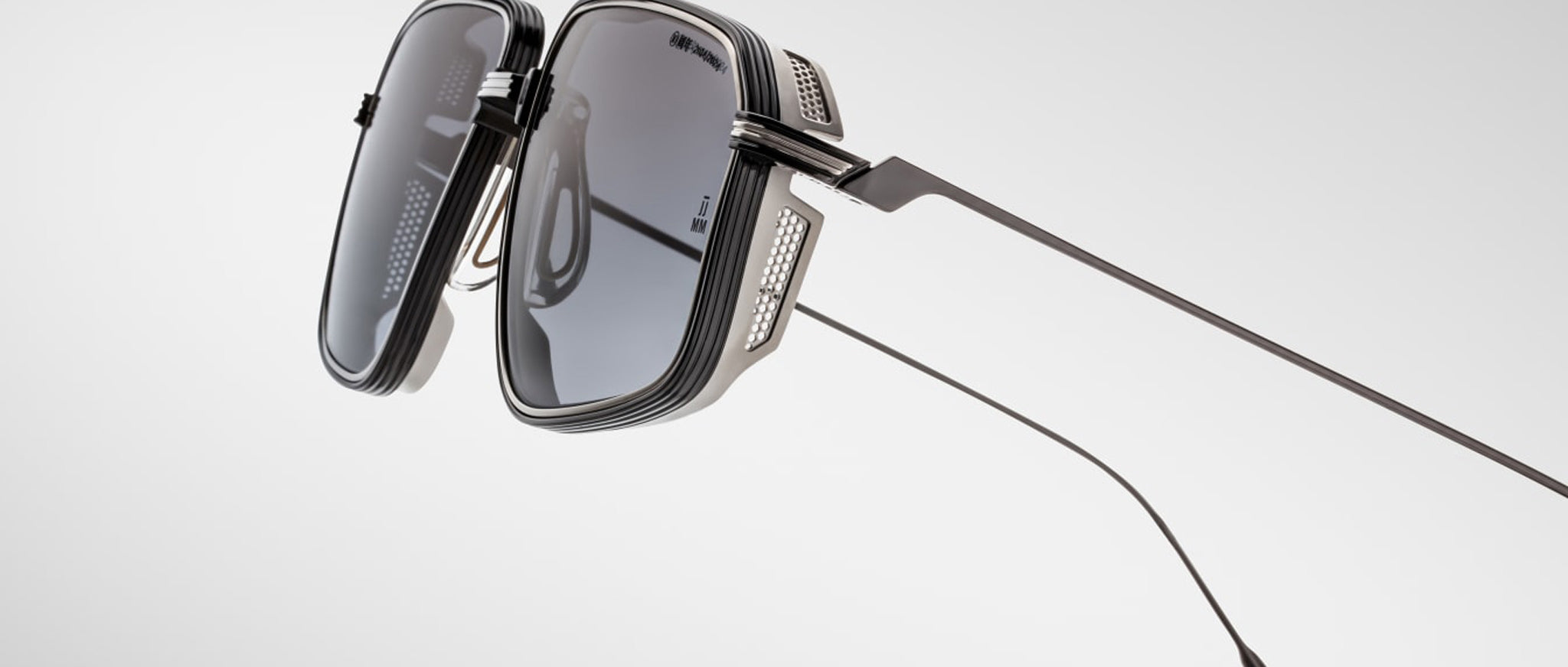 Close-up of Jacques Marie Mage Jagger sunglasses: futuristic, angular design with dark lenses, metal side shields, and thin beta titanium temples by Jacques Marie Mage, shown against a plain light background.