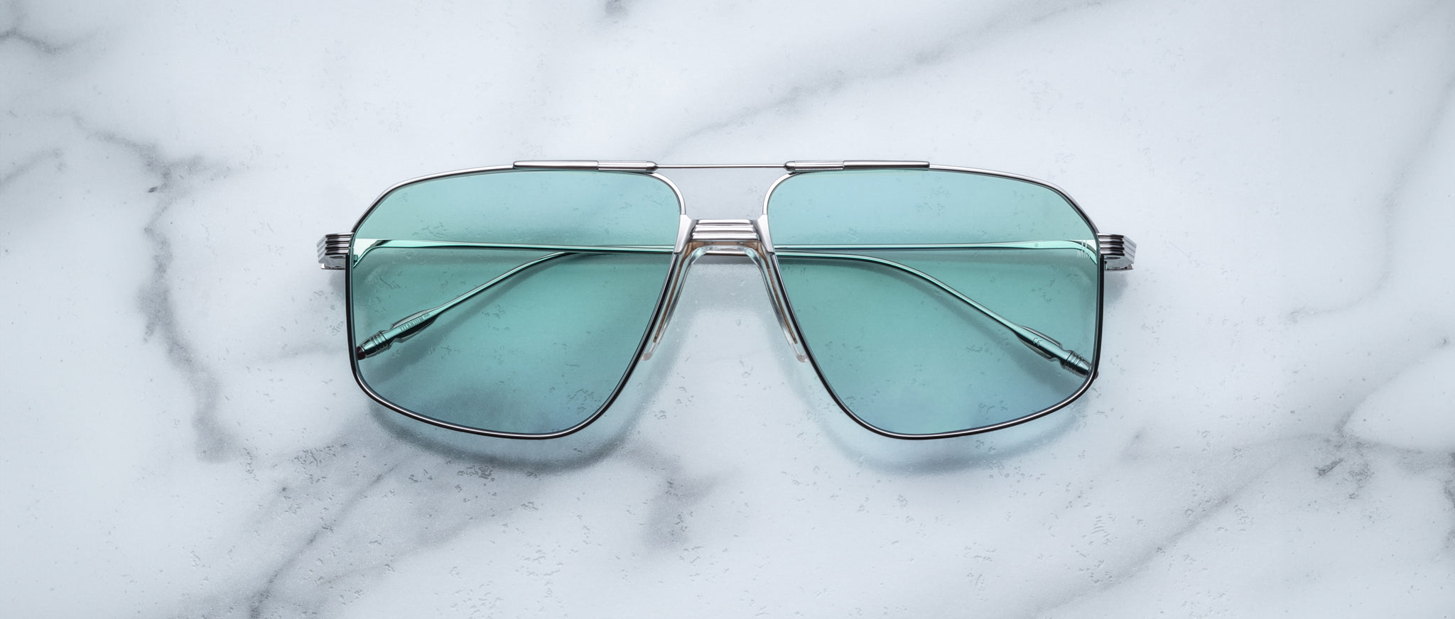 A pair of Jacques Marie Mage Jagger oversized aviator sunglasses with teal-tinted lenses and metal frames from Jacques Marie Mage rests on a white marble surface.