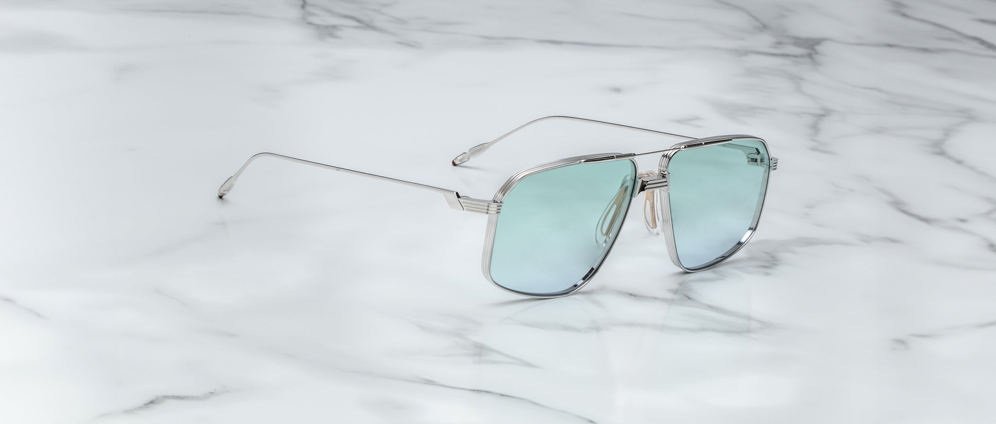 The Jacques Marie Mage Jagger sunglasses from Jacques Marie Mage feature an oversized aviator design with a silver metal frame, light blue reflective lenses, and beta titanium construction, shown elegantly on a white marble surface.