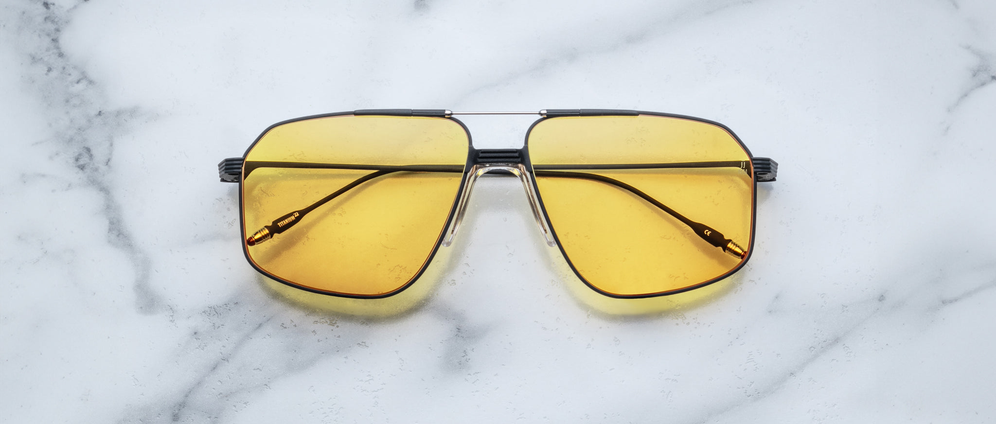 A pair of Jacques Marie Mage Jagger oversized aviator sunglasses with yellow-tinted lenses and sleek black arms rests on a white marble surface.