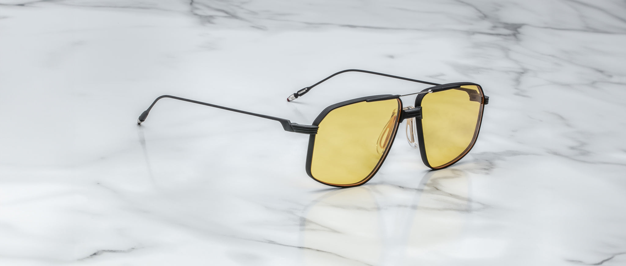 A pair of Jacques Marie Mage Jagger oversized aviator sunglasses with black frames and yellow lenses by Jacques Marie Mage sits on a white marble surface.