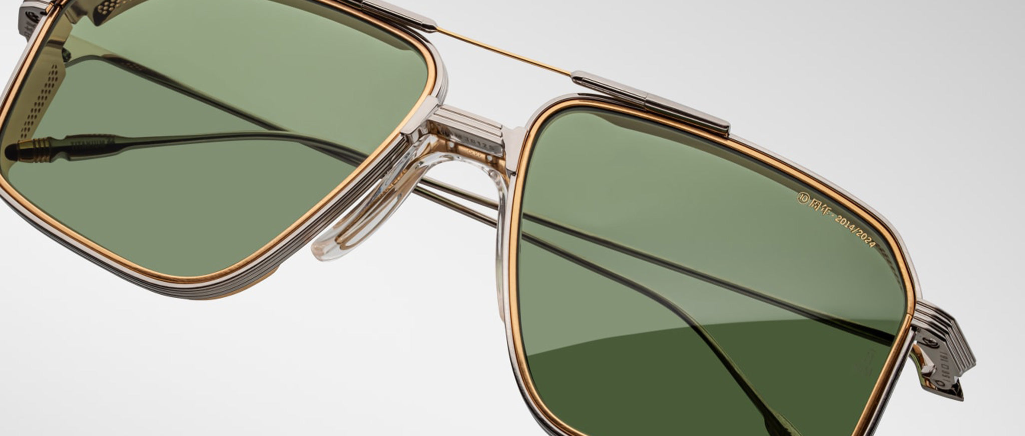 Close-up of Jacques Marie Mage Jagger sunglasses by Jacques Marie Mage, featuring green-tinted rectangular lenses, gold and silver beta titanium frames, and black earpieces against a plain background.