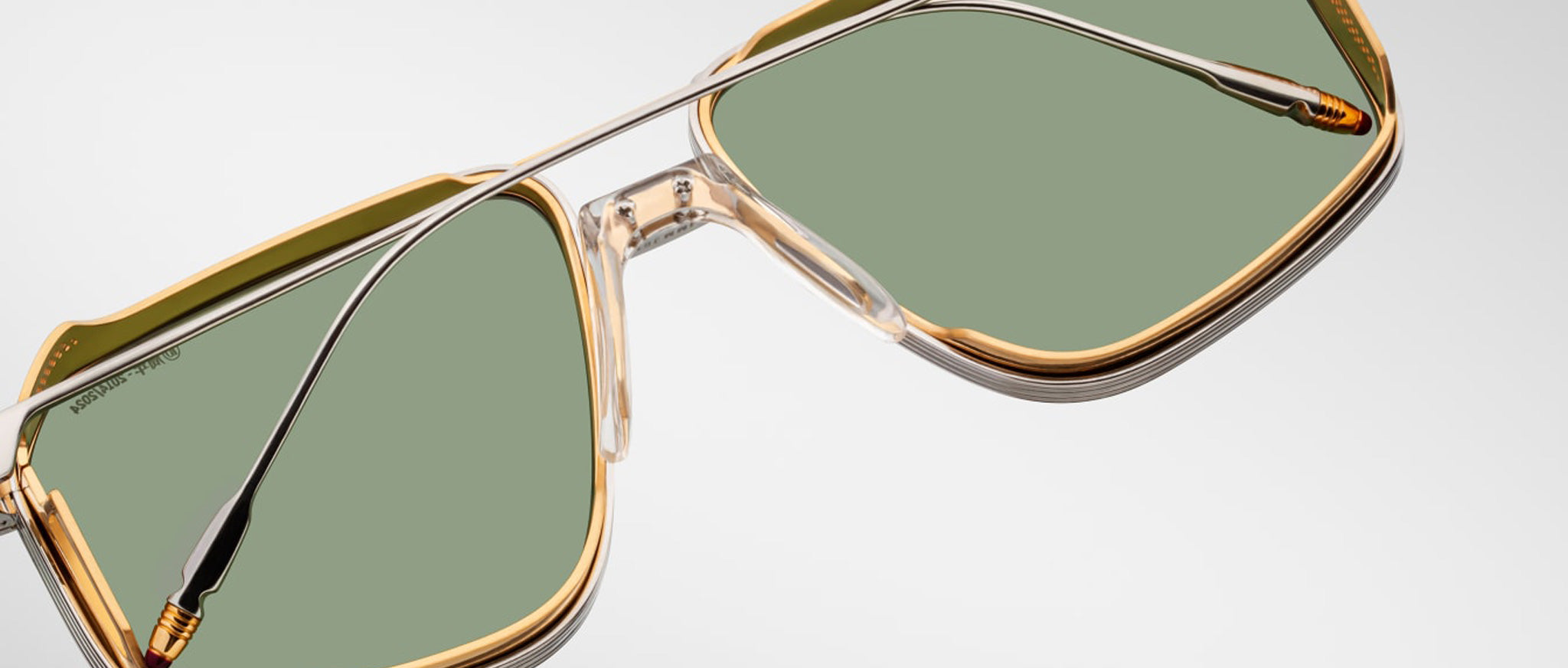 Close-up of Jacques Marie Mage Jagger aviator sunglasses by Jacques Marie Mage, featuring oversized transparent and gold frames, green-tinted lenses, and silver beta titanium arms, displayed on a light background.
