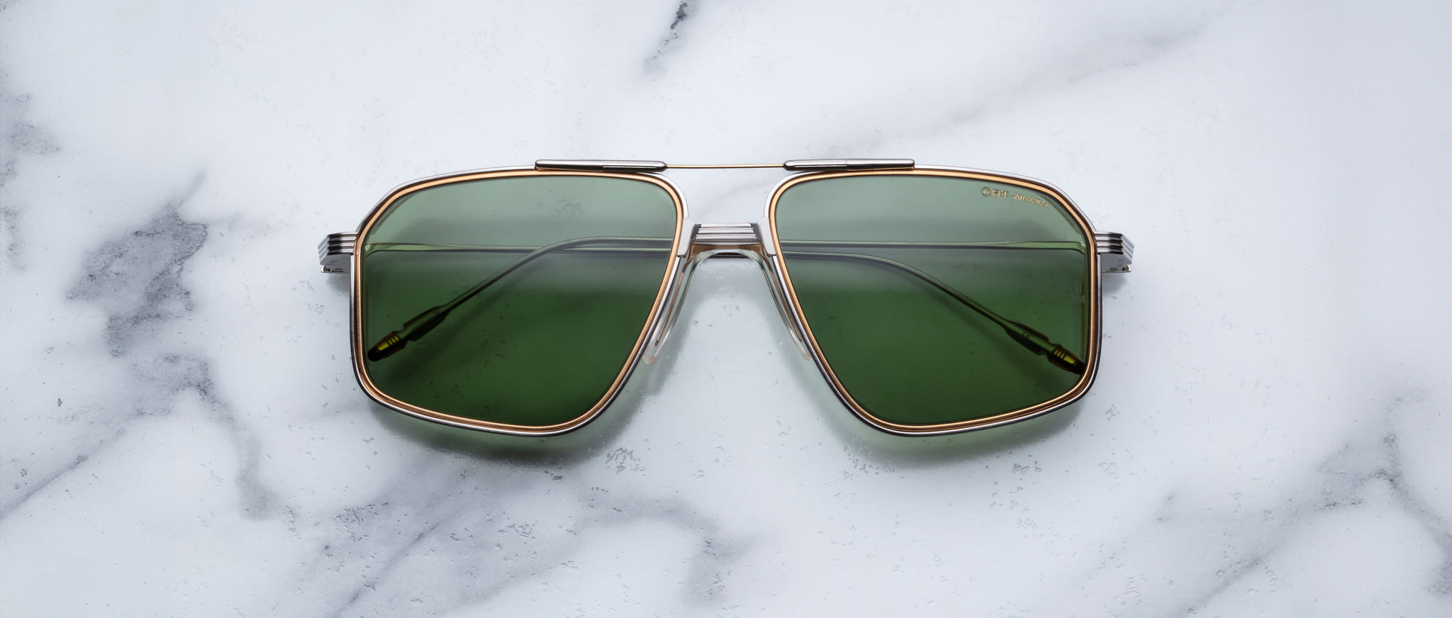 A pair of Jacques Marie Mage Jagger oversized aviator sunglasses with green lenses and gold beta titanium frames by Jacques Marie Mage sits on a white marble surface.