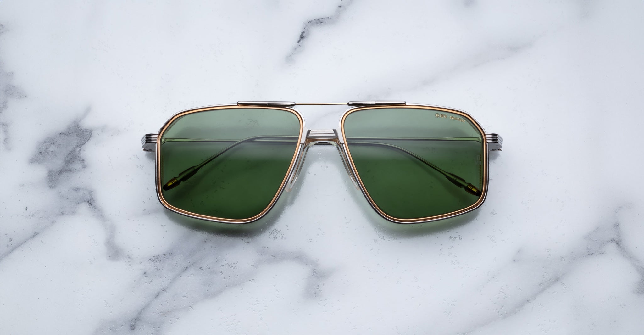 A pair of Jacques Marie Mage Jagger oversized aviator sunglasses with green lenses and gold beta titanium frames by Jacques Marie Mage sits on a white marble surface.