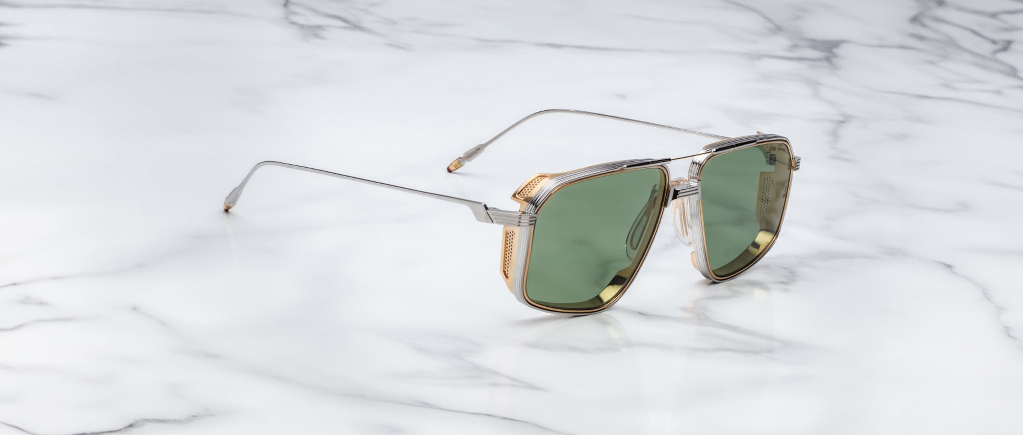 The Jacques Marie Mage Jagger sunglasses by Jacques Marie Mage feature oversized aviator frames with green lenses, gold accents, and a beta titanium metal frame, displayed stylishly on a white marble surface.