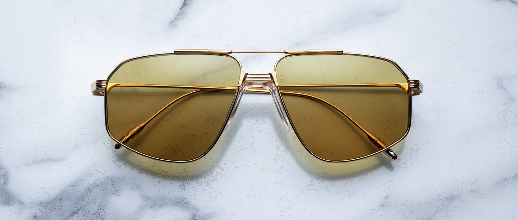 The Jagger sunglasses by Jacques Marie Mage, with oversized gold beta titanium frames and yellow-tinted lenses, are displayed on a white marble surface.