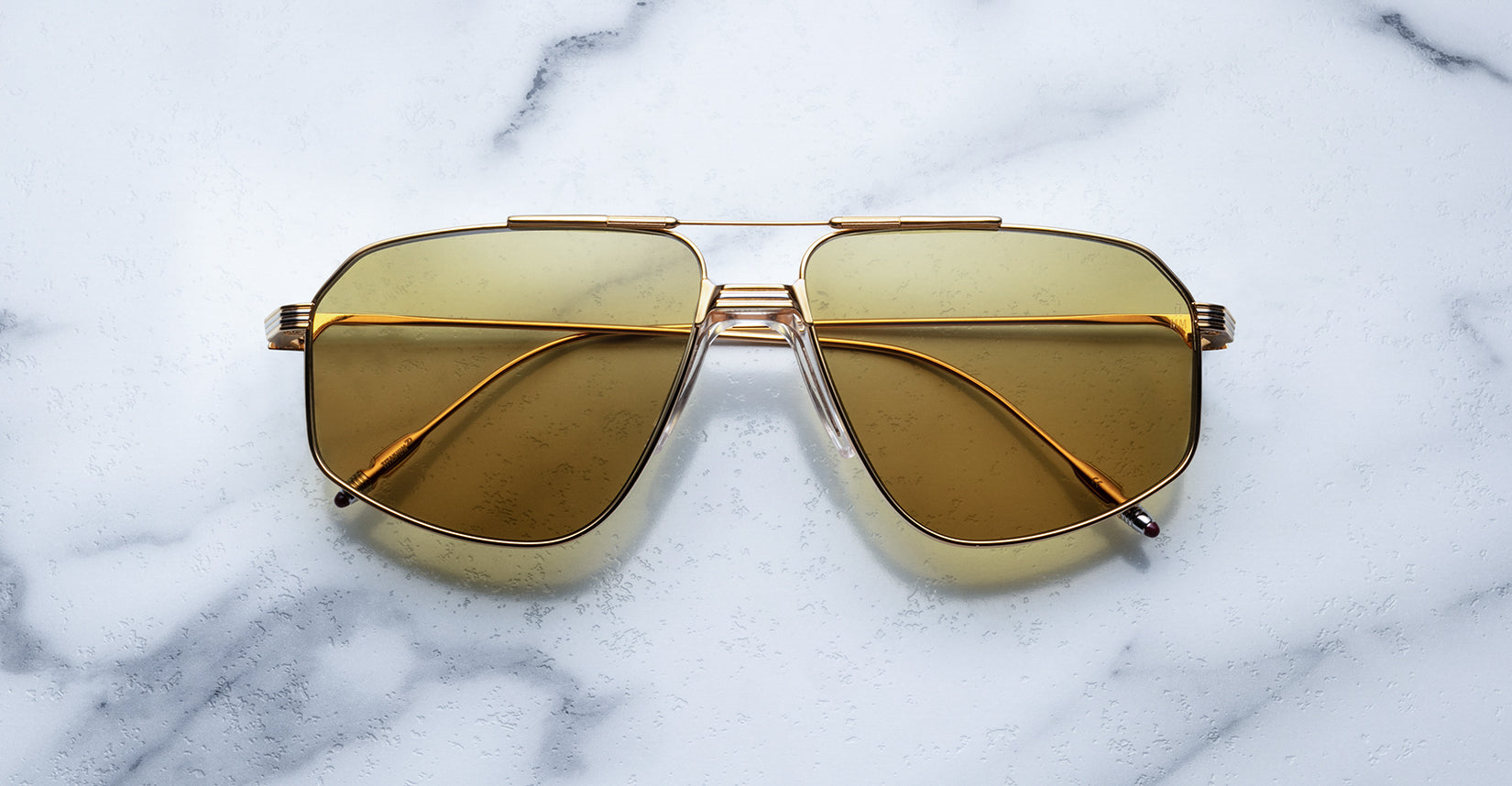 The Jagger sunglasses by Jacques Marie Mage, with oversized gold beta titanium frames and yellow-tinted lenses, are displayed on a white marble surface.
