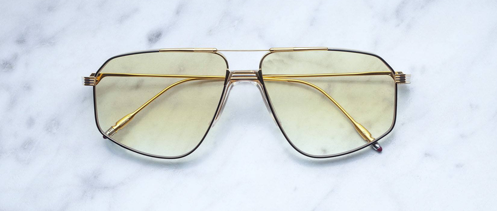 The Jacques Marie Mage Jagger sunglasses, featuring oversized gold aviator frames and yellow-tinted lenses, rest elegantly on a white marble surface.