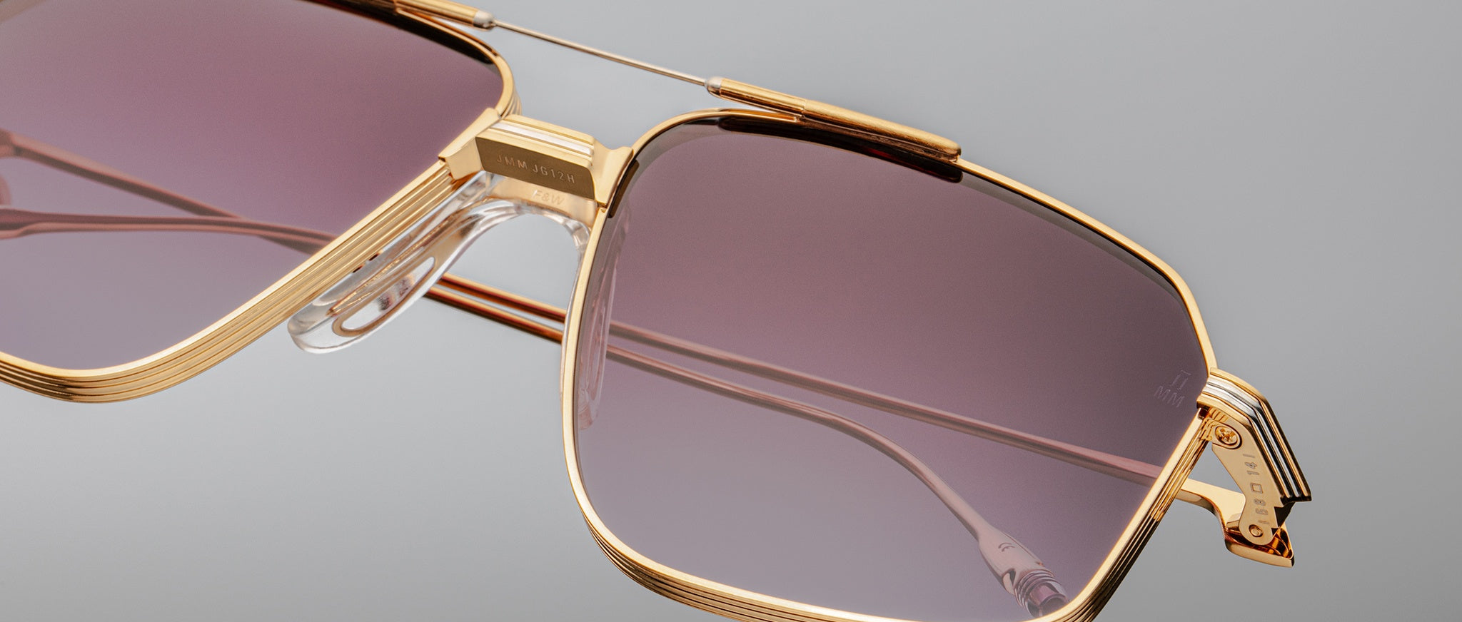 Close-up of oversized Jacques Marie Mage Jagger aviator sunglasses by Jacques Marie Mage, featuring gold beta titanium frames, purple-tinted lenses, clear nose pads, and detailed hinges, set against a plain gray background.