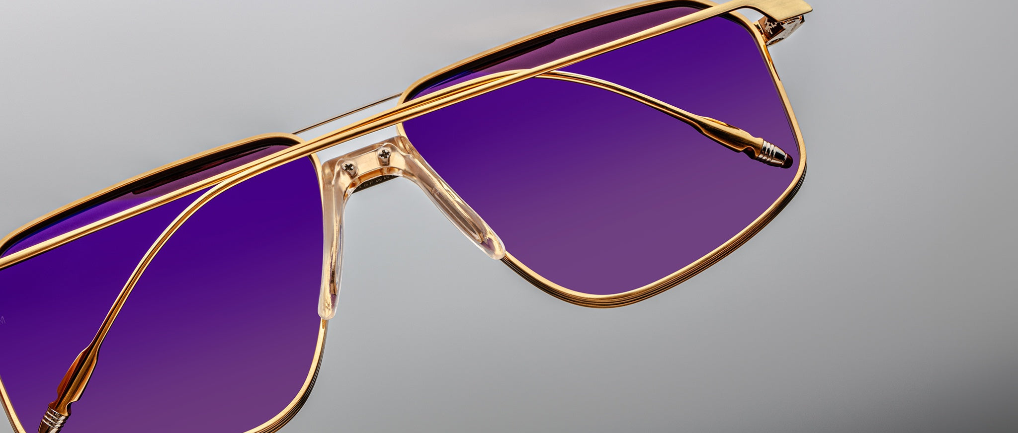 Close-up of Jacques Marie Mage Jagger oversized aviator sunglasses by Jacques Marie Mage, with gold beta titanium frames and purple-tinted lenses, shown on a gray background.