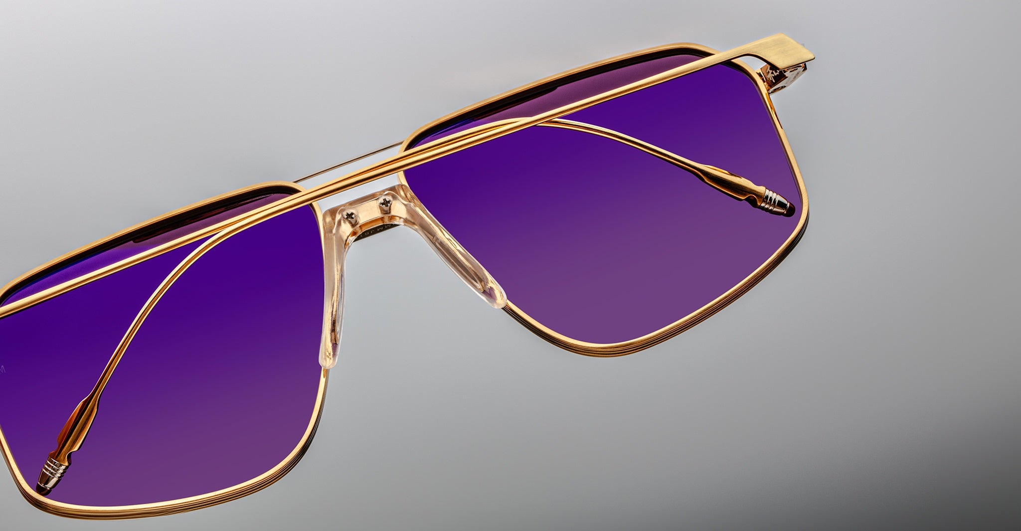 Close-up of Jacques Marie Mage Jagger oversized aviator sunglasses by Jacques Marie Mage, with gold beta titanium frames and purple-tinted lenses, shown on a gray background.
