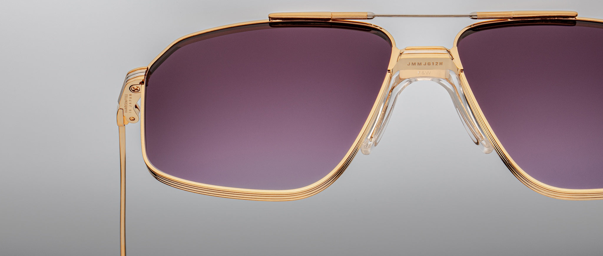 Close-up of Jacques Marie Mage Jagger oversized aviator sunglasses by Jacques Marie Mage, featuring gold frames and purple-tinted lenses, set against a plain light gray background.