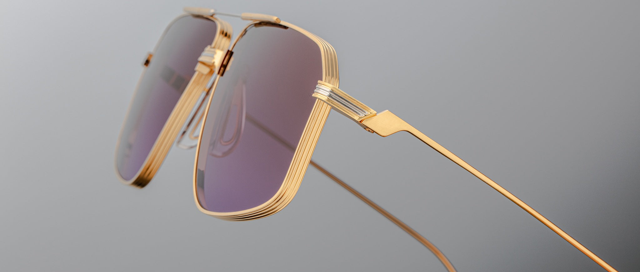 Close-up of Jacques Marie Mage Jagger oversized aviator sunglasses by Jacques Marie Mage, featuring gold frames and purple-tinted lenses on a plain gray background.