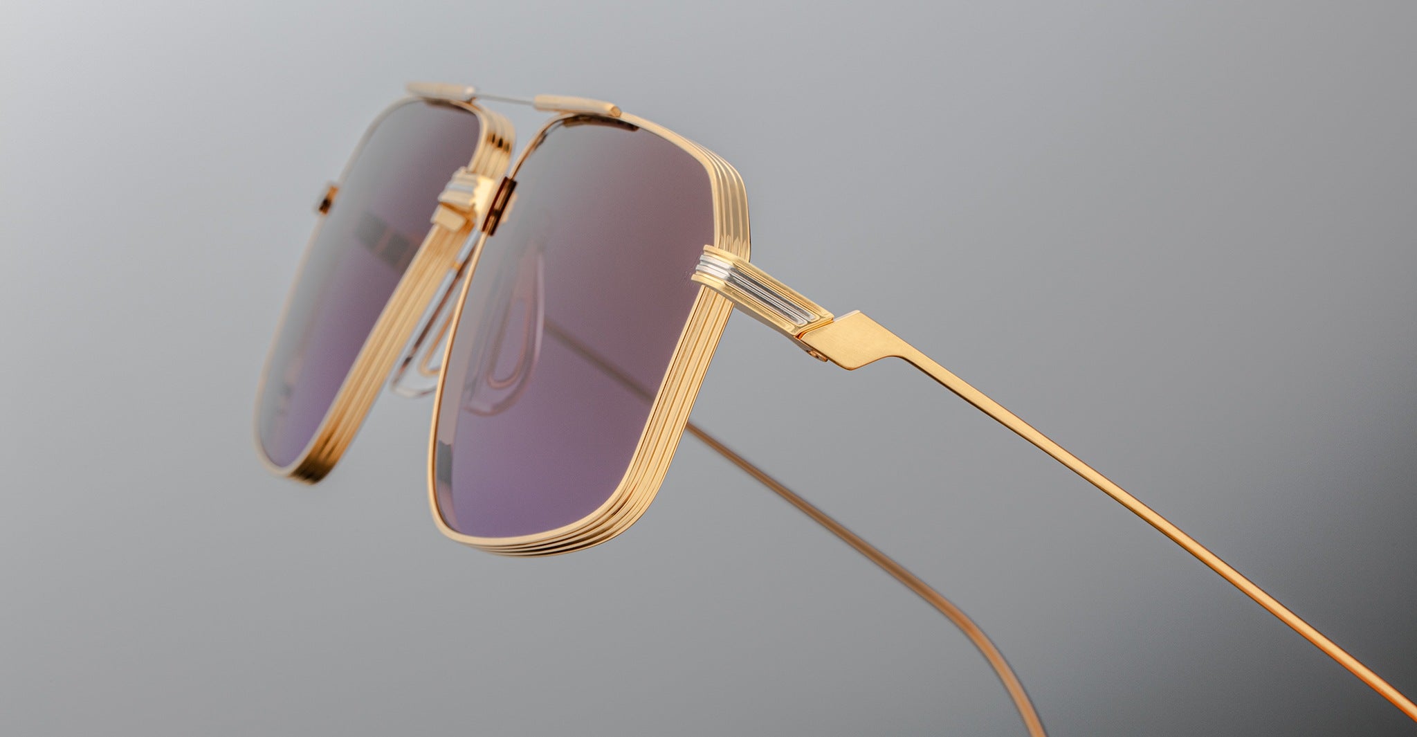 Close-up of Jacques Marie Mage Jagger oversized aviator sunglasses by Jacques Marie Mage, featuring gold frames and purple-tinted lenses on a plain gray background.