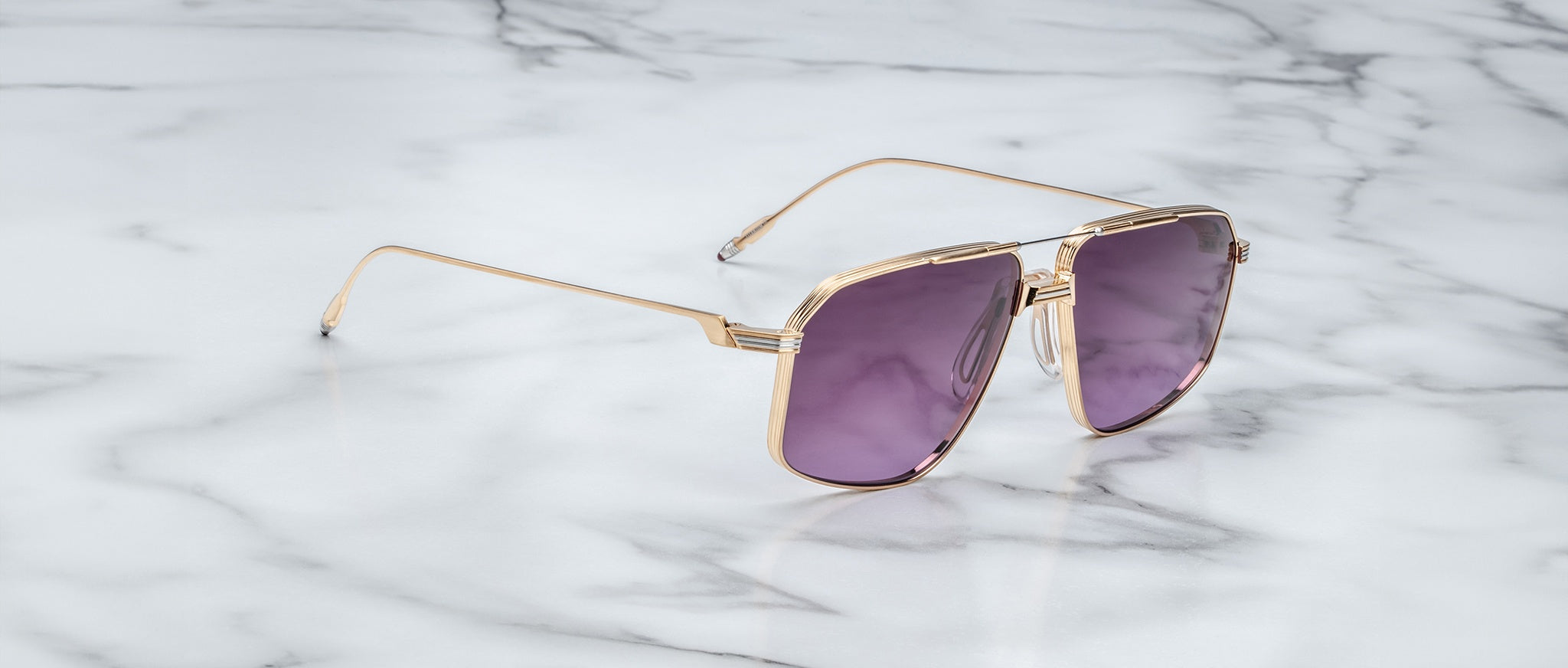 The Jacques Marie Mage Jagger sunglasses by Jacques Marie Mage, featuring gold frames and purple lenses, are displayed elegantly on a white marble surface.
