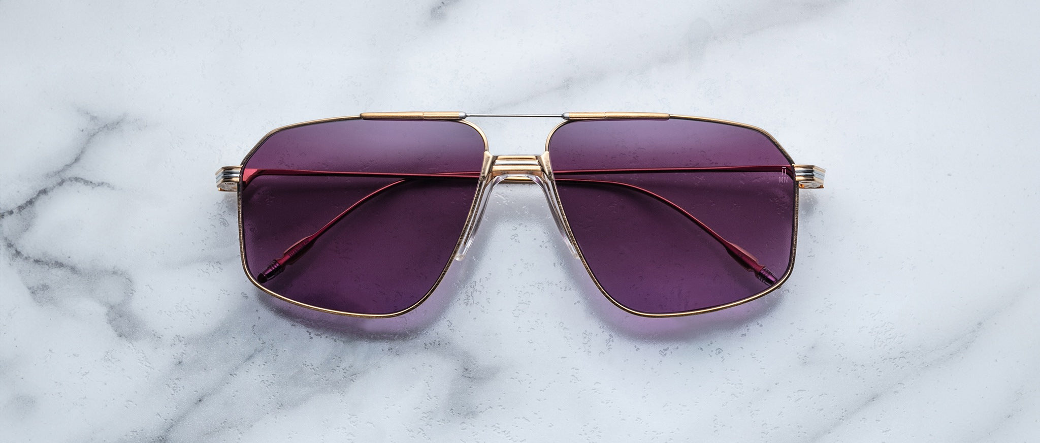 A pair of Jacques Marie Mage Jagger sunglasses with gold frames and purple lenses by Jacques Marie Mage sits on a white marble surface.