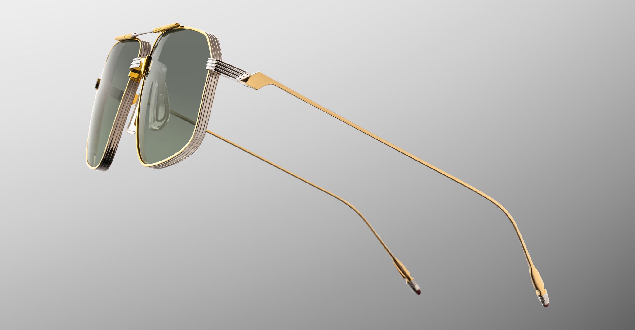 The Jacques Marie Mage Jagger by Jacques Marie Mage features gold-framed, rectangular sunglasses inspired by oversized aviators, crafted from beta titanium with green-tinted lenses, shown on a grey gradient background.