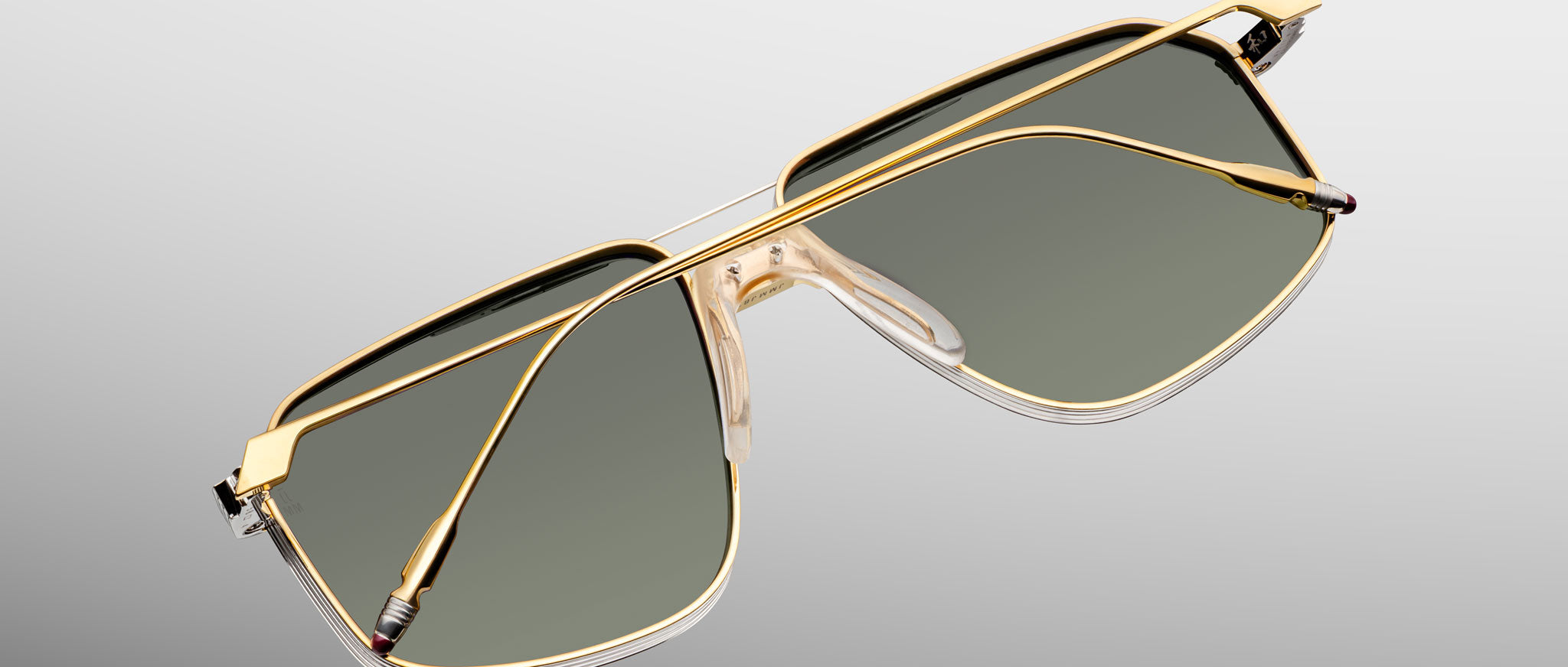 Jacques Marie Mage Jagger sunglasses by Jacques Marie Mage feature gold oversized aviator frames, dark green lenses, and clear nose pads, shown on a light gray background.